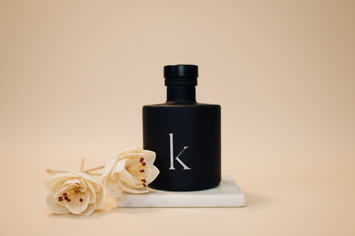 Black reed diffuser bottle with 'k' logo on a marble surface with flowers